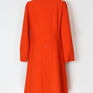 May include: A long, bright orange coat with a collar and a button closure. The coat has a fitted waist and a flared skirt.