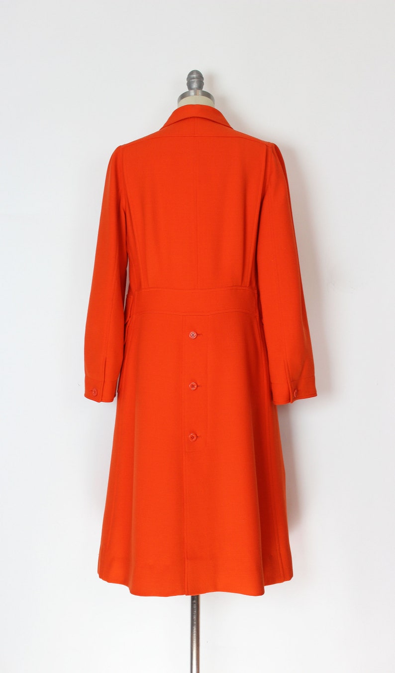 May include: A long, bright orange coat with a collar and a button closure. The coat has a fitted waist and a flared skirt.