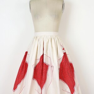 May include: A vintage-style skirt in cream with large red polka dot and geometric patterned panels. The skirt is displayed on a mannequin, showcasing its flared shape and design. The red patterns are outlined with a thin red border.
