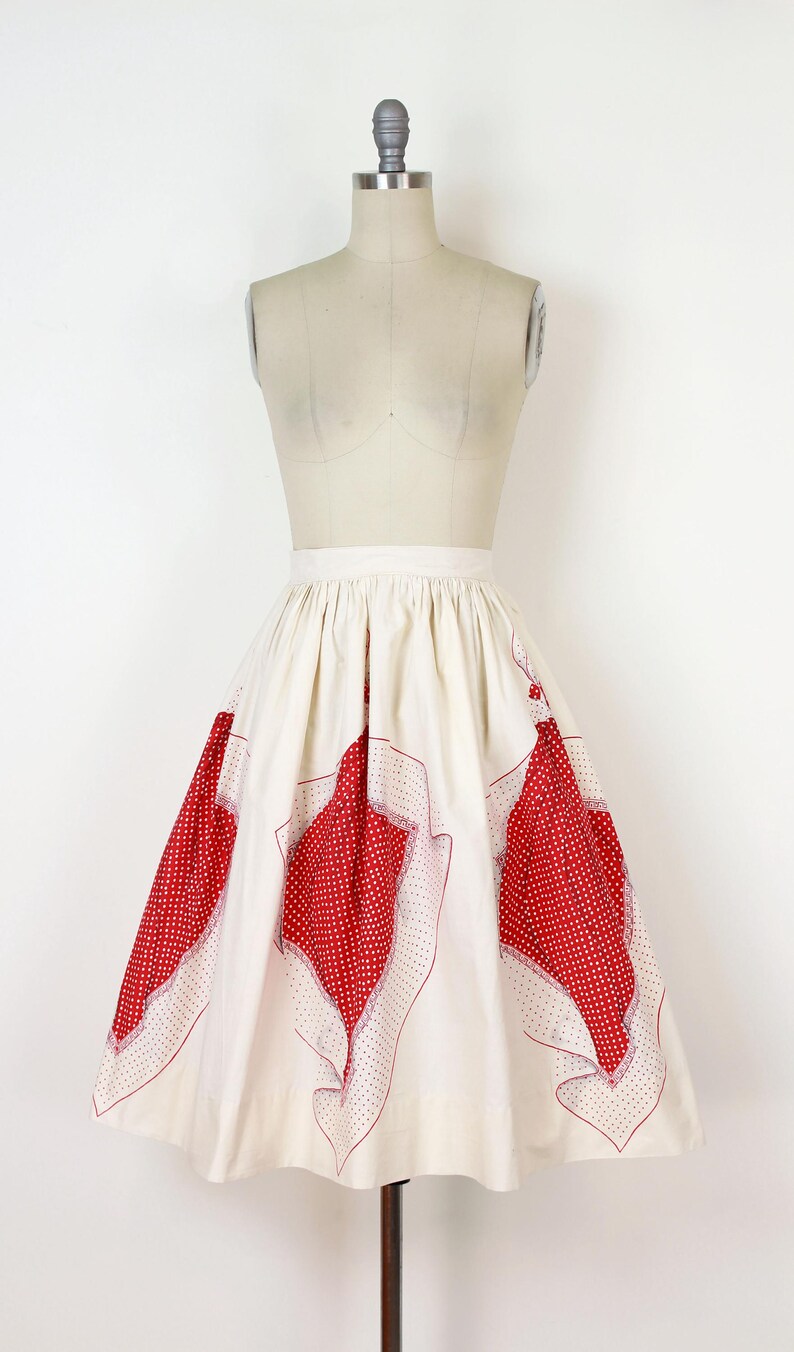 May include: A vintage-style skirt in cream with large red polka dot and geometric patterned panels. The skirt is displayed on a mannequin, showcasing its flared shape and design. The red patterns are outlined with a thin red border.