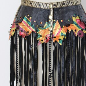 May include: A black leather skirt with a decorative waistband and fringe. The skirt features colorful geometric cutouts and is adorned with black beads and gold-colored buttons. The fringe hangs vertically, adding movement to the design.