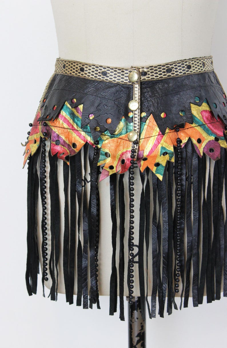 May include: A black leather skirt with a decorative waistband and fringe. The skirt features colorful geometric cutouts and is adorned with black beads and gold-colored buttons. The fringe hangs vertically, adding movement to the design.