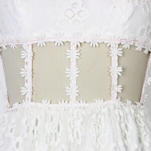 May include: A white lace dress with a floral design. The dress has a sheer panel at the waist, trimmed with daisy-shaped lace. The bodice and skirt are made of intricate lace, creating a romantic and elegant look.