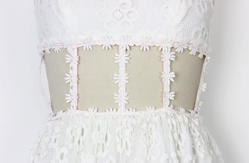 May include: A white lace dress with a floral design. The dress has a sheer panel at the waist, trimmed with daisy-shaped lace. The bodice and skirt are made of intricate lace, creating a romantic and elegant look.