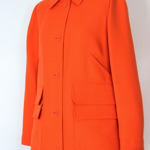 May include: A long, bright orange coat with a button-up front and large patch pockets. The coat has a classic, tailored look.
