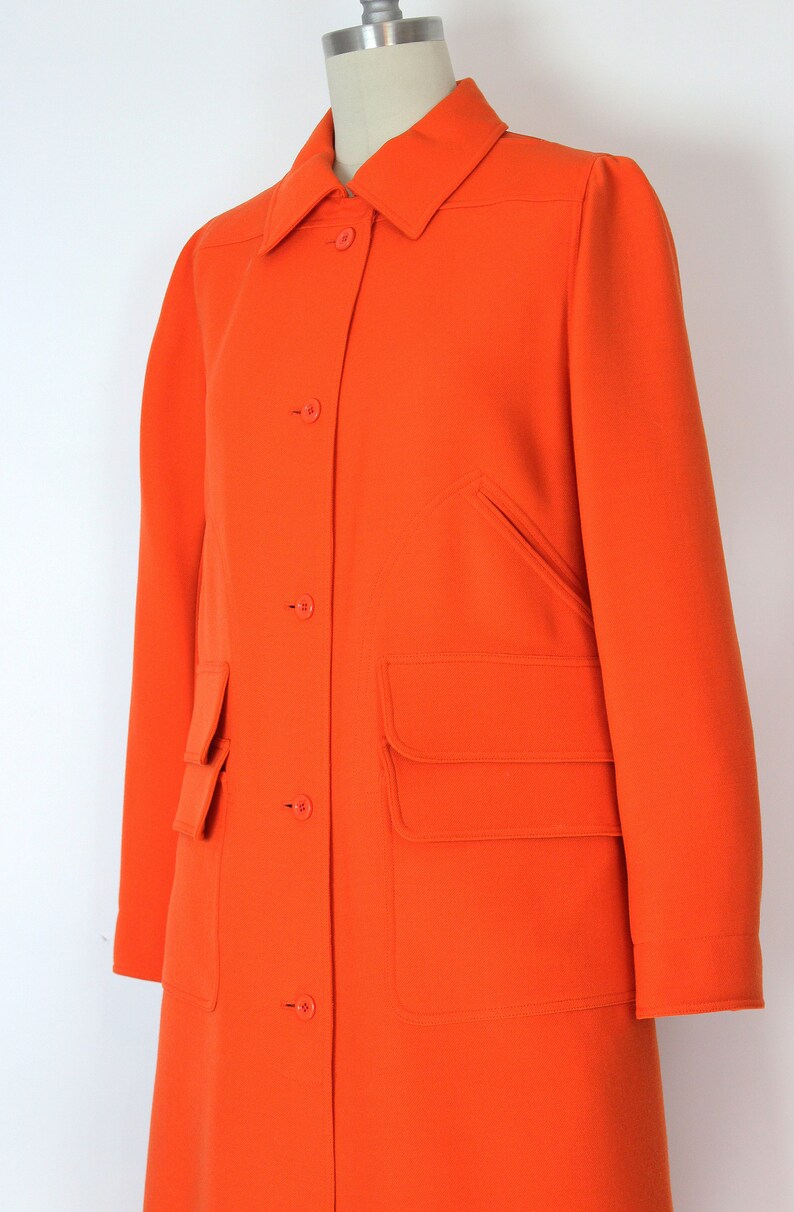 May include: A long, bright orange coat with a button-up front and large patch pockets. The coat has a classic, tailored look.