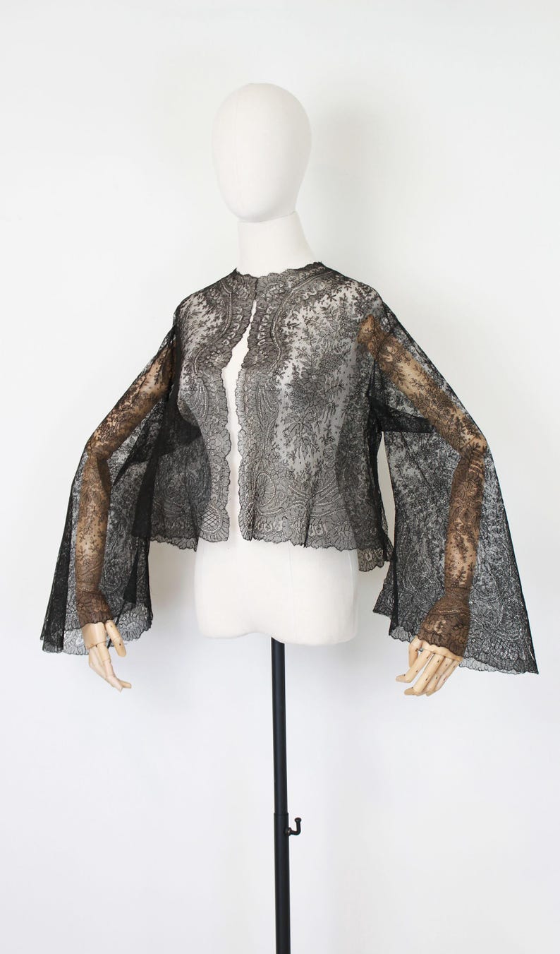 May include: Black lace jacket with flared sleeves, displayed on a mannequin. The jacket features an open front and intricate floral patterns. The sleeves extend to the wrists, showcasing a delicate design.
