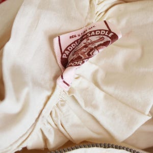 May include: Close-up of a cream-colored garment with a zipper and a pink label. The label reads "PADDLE PORT SADDLE" with a graphic of a person in a canoe. The fabric has a textured appearance.