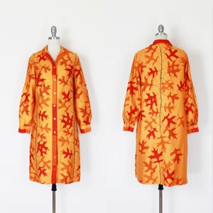 May include: A long-sleeved button-down shirt dress in a vibrant orange and yellow hue, featuring a repeating pattern of red and orange oak leaves. The garment includes a pointed collar, a central button placket, and cuffed sleeves.