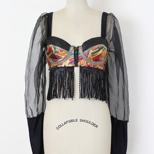 May include: A black bustier top with a colorful, geometric design on the bodice. The top features a zipper closure, fringe, and sheer black sleeves. The words "COLLAPSIBLE SHOULDER" are printed on the mannequin.