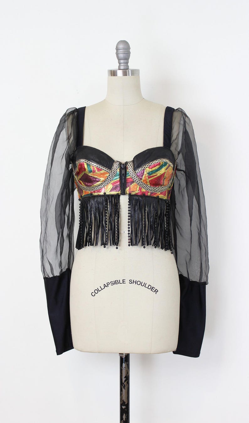 May include: A black bustier top with a colorful, geometric design on the bodice. The top features a zipper closure, fringe, and sheer black sleeves. The words "COLLAPSIBLE SHOULDER" are printed on the mannequin.