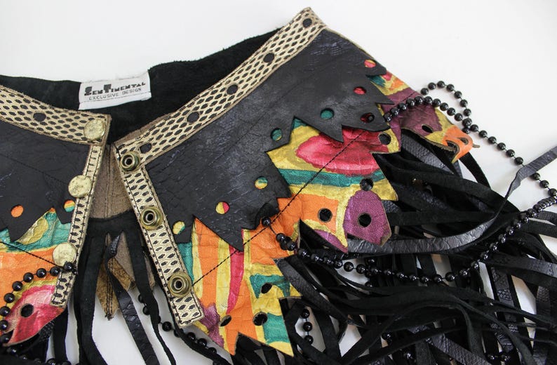 May include: A black leather vest with colorful, abstract painted accents and fringe. The vest features a decorative trim with gold-colored buttons and a beaded necklace. The label reads "SENTIMENTAL EXCLUSIVE DESIGN."