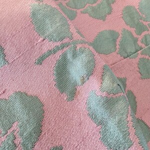 May include: Close-up of a pink fabric with a floral pattern in a muted teal color. The design features large, stylized rose blooms and leaves, creating a textured, woven appearance. The fabric is folded, showing the pattern's repeat.