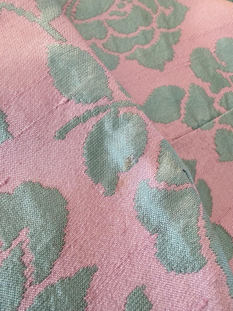 May include: Close-up of a pink fabric with a floral pattern in a muted teal color. The design features large, stylized rose blooms and leaves, creating a textured, woven appearance. The fabric is folded, showing the pattern's repeat.