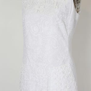 May include: White sleeveless dress with a round neckline and intricate floral embroidery. The dress features a subtle all-over paisley pattern and decorative trim along the neckline and hem. The dress is displayed on a mannequin.