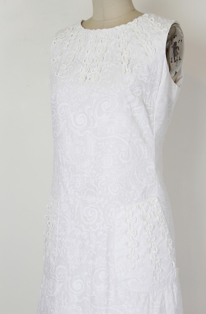 May include: White sleeveless dress with a round neckline and intricate floral embroidery. The dress features a subtle all-over paisley pattern and decorative trim along the neckline and hem. The dress is displayed on a mannequin.
