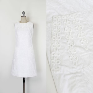May include: A sleeveless white dress with a textured floral pattern. The dress is displayed on a mannequin. A close-up shows a decorative trim with a looped design.