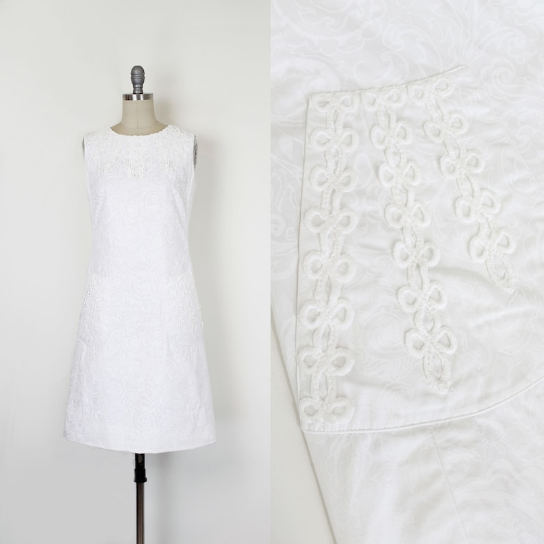 May include: A sleeveless white dress with a textured floral pattern. The dress is displayed on a mannequin. A close-up shows a decorative trim with a looped design.