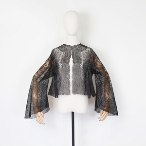 May include: A black lace jacket with long, flared sleeves is displayed on a mannequin. The jacket features a delicate floral pattern and scalloped edges. The sheer fabric allows a glimpse of the mannequin's form beneath.