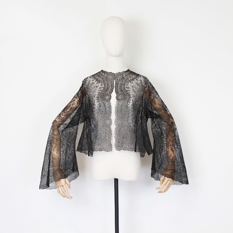 May include: A black lace jacket with long, flared sleeves is displayed on a mannequin. The jacket features a delicate floral pattern and scalloped edges. The sheer fabric allows a glimpse of the mannequin's form beneath.