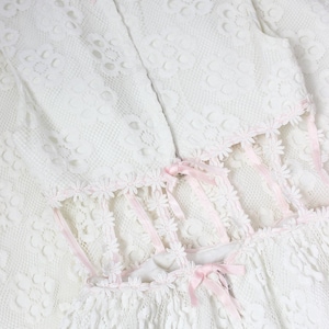 May include: A white lace dress with floral detailing and pink ribbon trim. The dress has a high neckline, fitted bodice, and a flared skirt. Pink bows and floral edging add a delicate touch to this vintage-style garment.