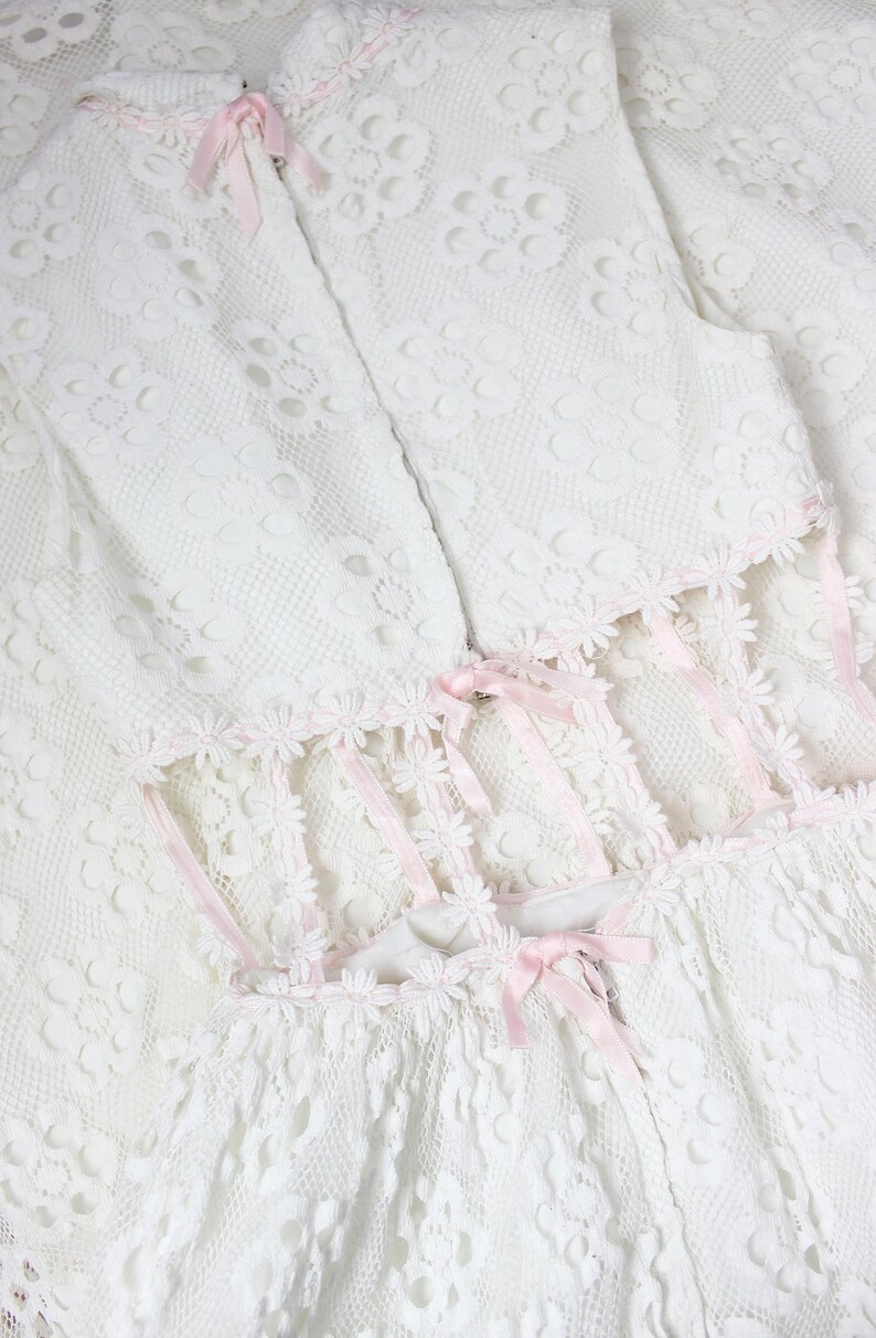 May include: A white lace dress with floral detailing and pink ribbon trim. The dress has a high neckline, fitted bodice, and a flared skirt. Pink bows and floral edging add a delicate touch to this vintage-style garment.