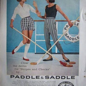 May include: Vintage advertisement featuring two women playing a game with wooden mallets and rings. One woman wears a white shirt and plaid shorts, the other a black top and striped pants. The text reads "Paddle & Saddle Sportswear."