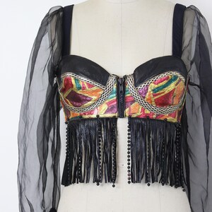 May include: A black bustier top with a colorful geometric pattern and a zipper closure. The top features black fringe and sheer black sleeves. The text "COLLAPSIBLE SHOULDER" is printed on the mannequin.