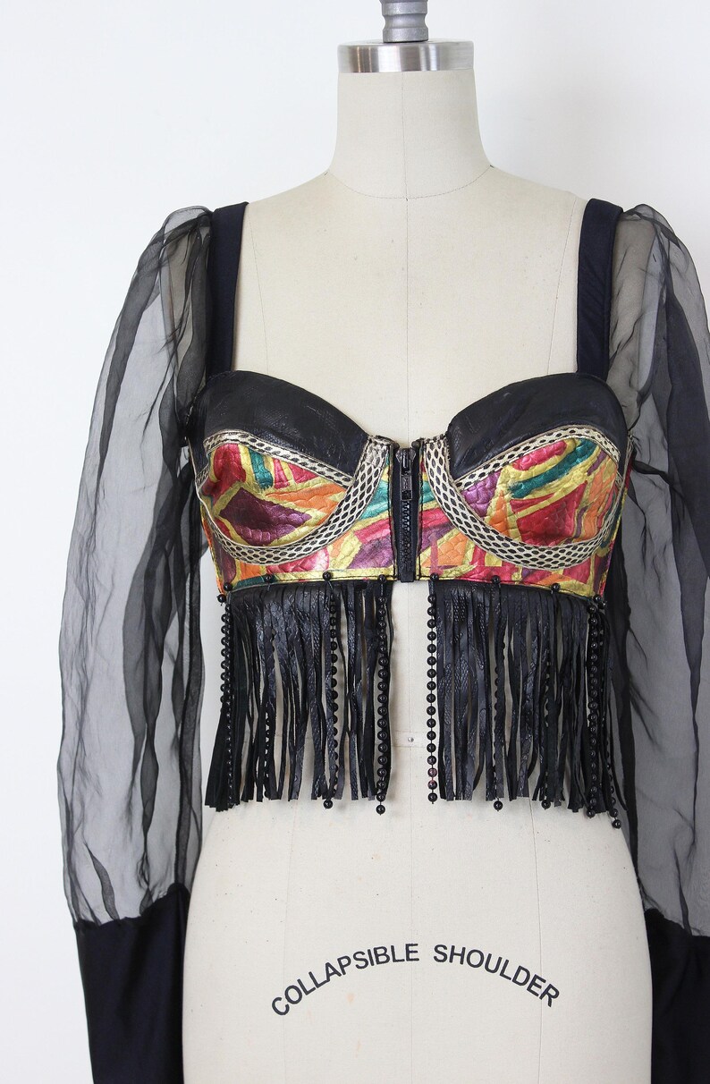 May include: A black bustier top with a colorful geometric pattern and a zipper closure. The top features black fringe and sheer black sleeves. The text "COLLAPSIBLE SHOULDER" is printed on the mannequin.