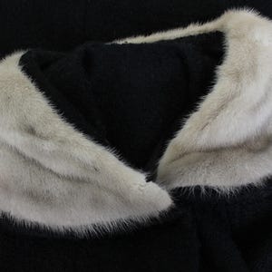 Puede incluir: Primer plano de un abrigo negro con cuello de piel gris claro. El cuello de piel es grueso y esponjoso, contrastando con la tela oscura del abrigo. El cuello es una caracter&iacute;stica destacada de la prenda.