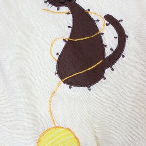May include: A white knitted sweater with a brown cat and yellow yarn ball embroidered on the front.