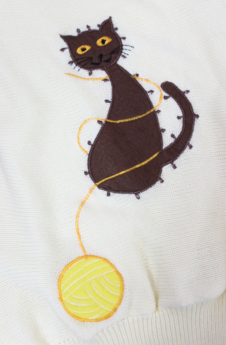 May include: A white knitted sweater with a brown cat and yellow yarn ball embroidered on the front.