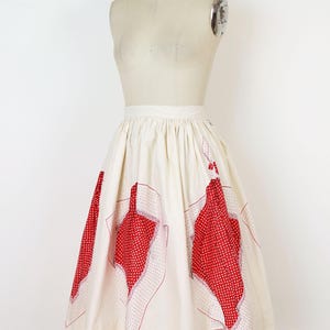 May include: Cream-colored vintage skirt with red polka dot and floral-shaped accents. The skirt is displayed on a mannequin. The red accents are outlined with a dotted pattern. The skirt has a high waist and a flared silhouette.