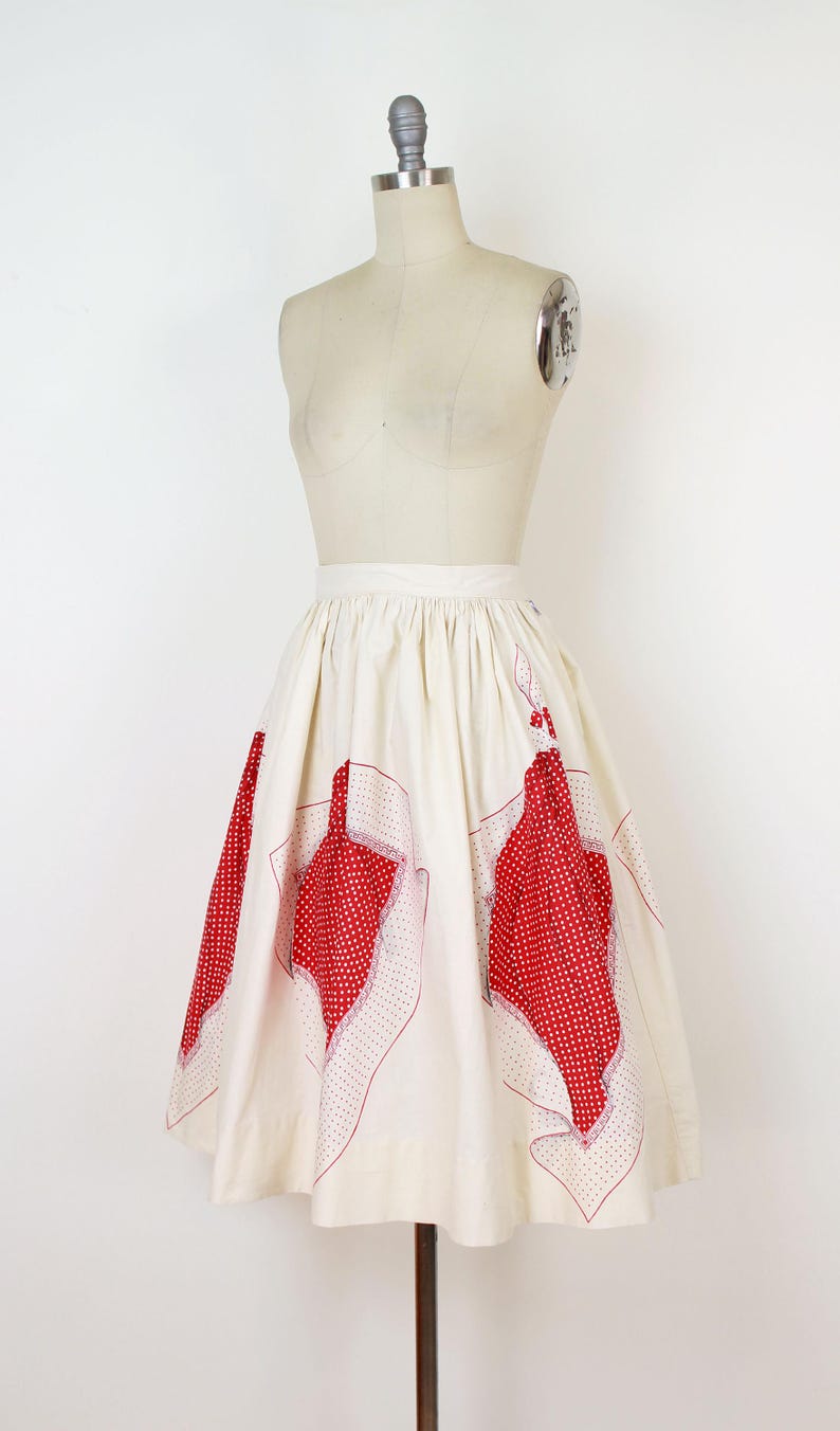 May include: Cream-colored vintage skirt with red polka dot and floral-shaped accents. The skirt is displayed on a mannequin. The red accents are outlined with a dotted pattern. The skirt has a high waist and a flared silhouette.