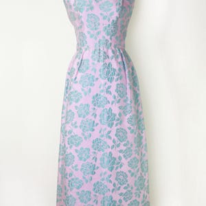 May include: A sleeveless, knee-length dress in a light pink hue, adorned with a repeating pattern of teal-colored floral designs. The dress features a fitted bodice and a slightly flared skirt, displayed on a mannequin.