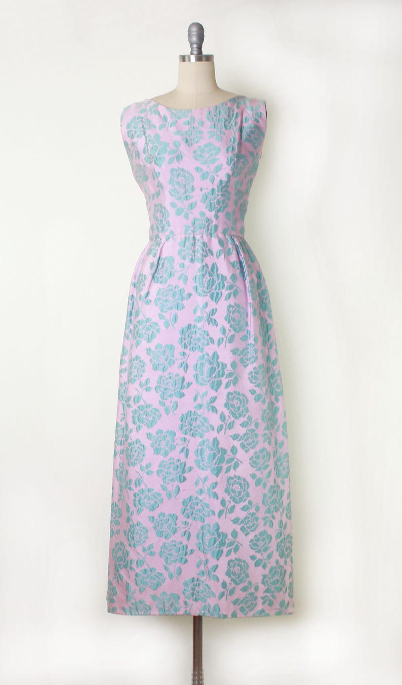 May include: A sleeveless, knee-length dress in a light pink hue, adorned with a repeating pattern of teal-colored floral designs. The dress features a fitted bodice and a slightly flared skirt, displayed on a mannequin.