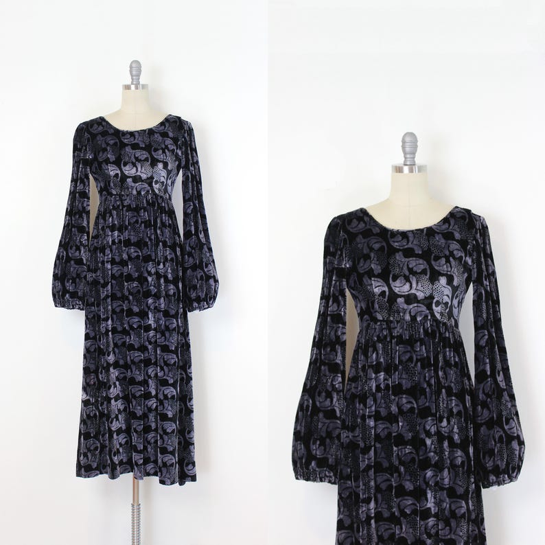 May include: A long, dark gray velvet dress with a repeating skull pattern. The dress features a round neckline, long sleeves with a balloon shape, and a gathered waist. The dress is displayed on a mannequin.