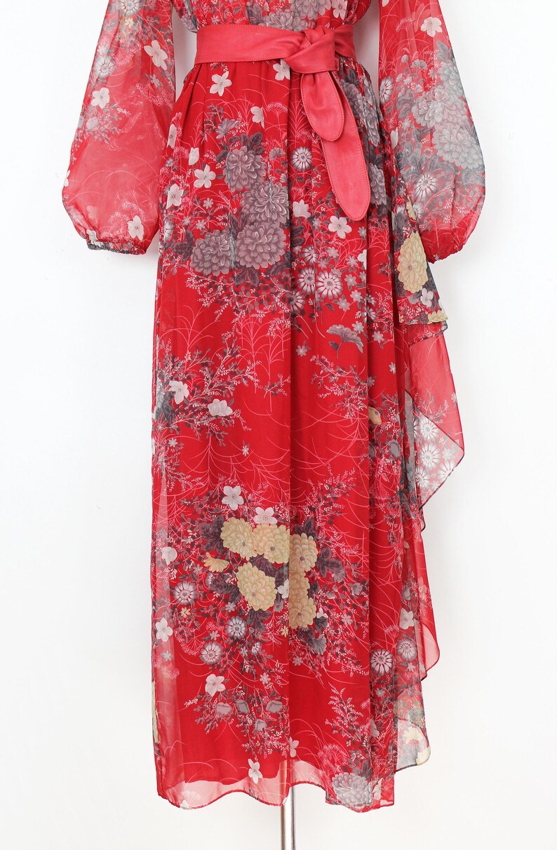 May include: A red maxi dress with a floral print and a ruffled hem. The dress has a V-neckline and long sleeves with a gathered detail at the shoulders. The dress is cinched at the waist with a matching red sash.