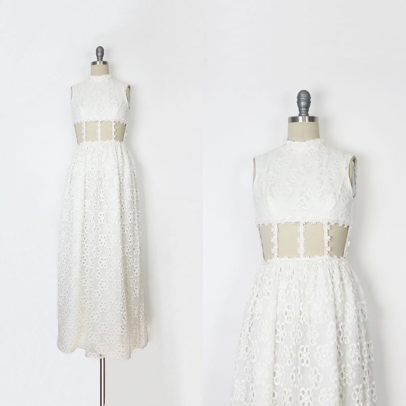 May include: A white lace dress with a high neckline and a sheer panel at the waist. The dress is sleeveless and has a full skirt. The dress is displayed on a mannequin.