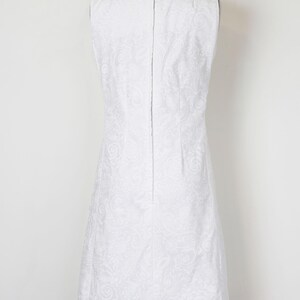 May include: A sleeveless white dress with a subtle floral pattern, displayed on a mannequin. The dress has a simple, elegant design with a straight silhouette and a zipper down the back. The fabric appears to have a slight sheen.