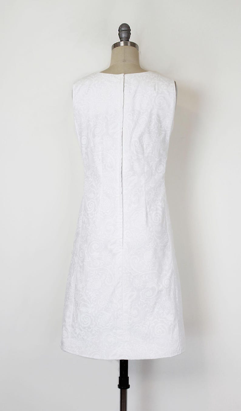 May include: A sleeveless white dress with a subtle floral pattern, displayed on a mannequin. The dress has a simple, elegant design with a straight silhouette and a zipper down the back. The fabric appears to have a slight sheen.