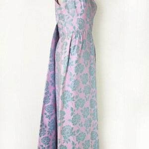 May include: A sleeveless, floor-length dress on a mannequin. The dress features a pink background with a teal floral pattern. The dress has a flowing skirt and a matching fabric detail that drapes from the back.