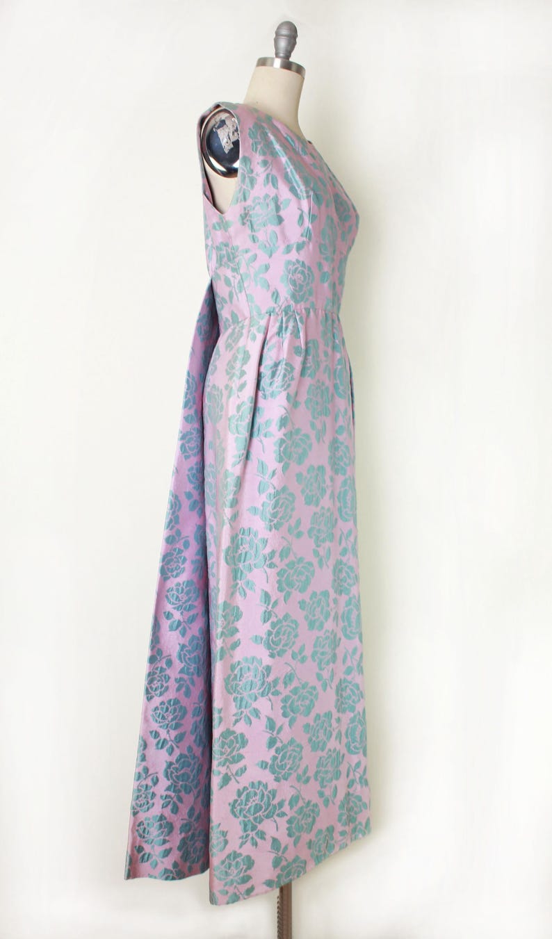 May include: A sleeveless, floor-length dress on a mannequin. The dress features a pink background with a teal floral pattern. The dress has a flowing skirt and a matching fabric detail that drapes from the back.