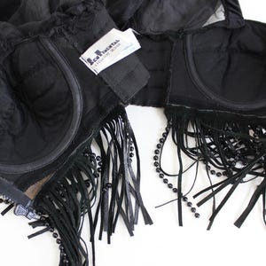 May include: Black lingerie set featuring a bra with fringe and beaded accents. The bra has a zipper closure and a tag that reads "L'Agent by Agent Provocateur". The set is made of sheer and opaque black fabric.
