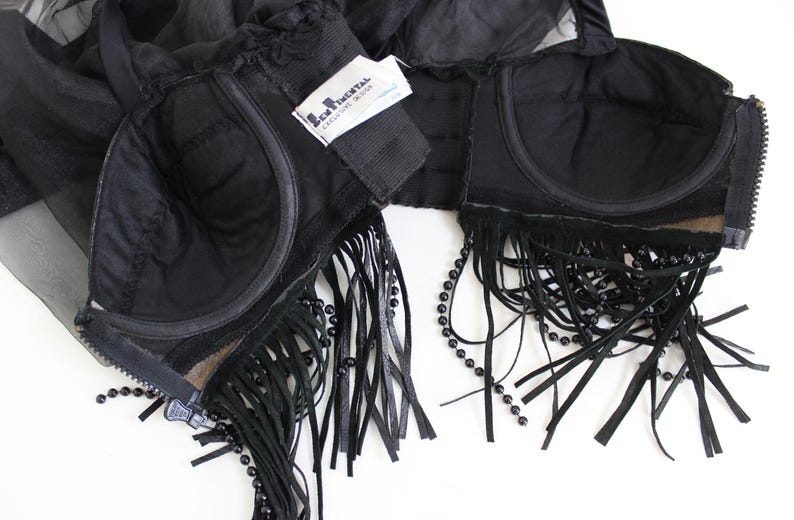 May include: Black lingerie set featuring a bra with fringe and beaded accents. The bra has a zipper closure and a tag that reads "L'Agent by Agent Provocateur". The set is made of sheer and opaque black fabric.