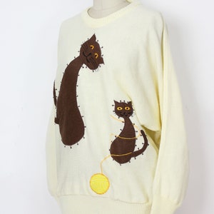 May include: A cream-colored knitted sweater with brown cat appliques and a yellow yarn ball. The cats are embroidered with brown yarn and have orange eyes.