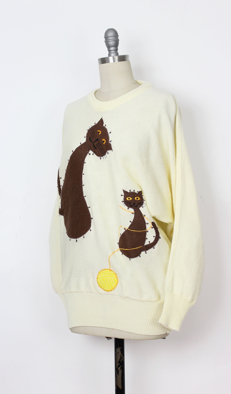 May include: A cream-colored knitted sweater with brown cat appliques and a yellow yarn ball. The cats are embroidered with brown yarn and have orange eyes.