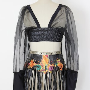 May include: Black crop top with sheer sleeves and a matching skirt. The skirt features a colorful, patterned design with fringe and beaded accents. The outfit is displayed on a mannequin.