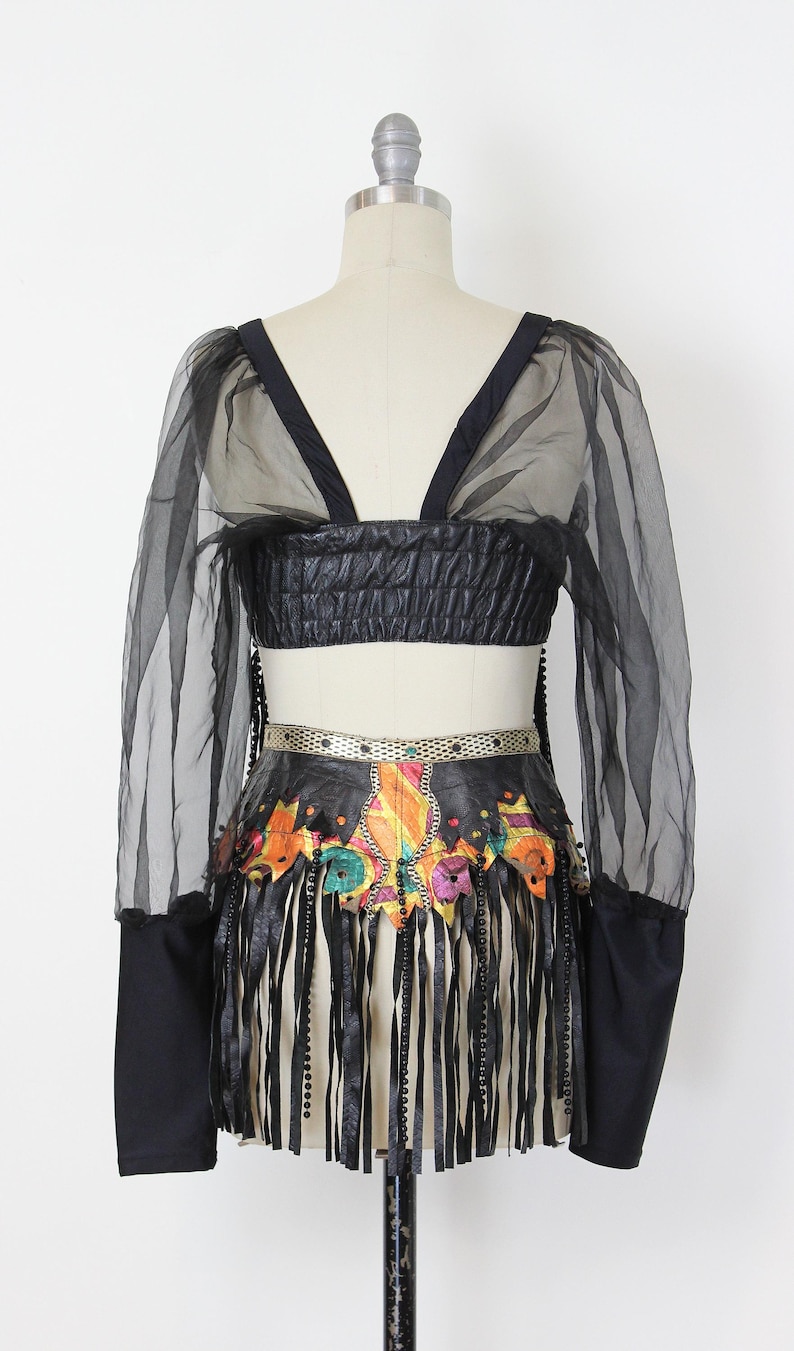 May include: Black crop top with sheer sleeves and a matching skirt. The skirt features a colorful, patterned design with fringe and beaded accents. The outfit is displayed on a mannequin.