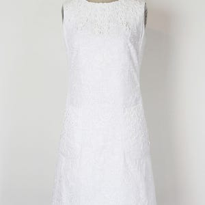 May include: A sleeveless white dress with an intricate floral pattern and eyelet detailing. The dress is displayed on a mannequin, showcasing its A-line silhouette and round neckline. The fabric appears to be cotton or a similar lightweight material.